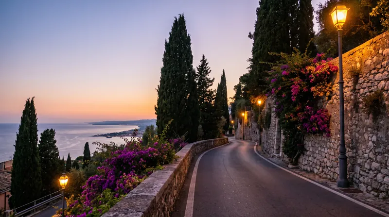 A narrow hillside road lined with cypress trees and stone walls above the sea in eastern Sicily, at dusk