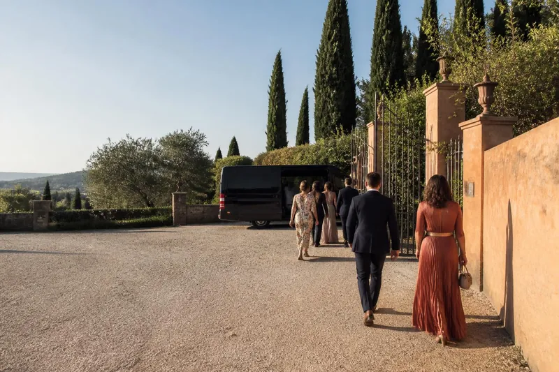 Wedding guests in formal clothes walking from a villa gate toward a black minibus